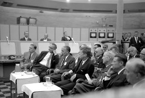 JFK and LBJ with others sitting on chairs in a room.