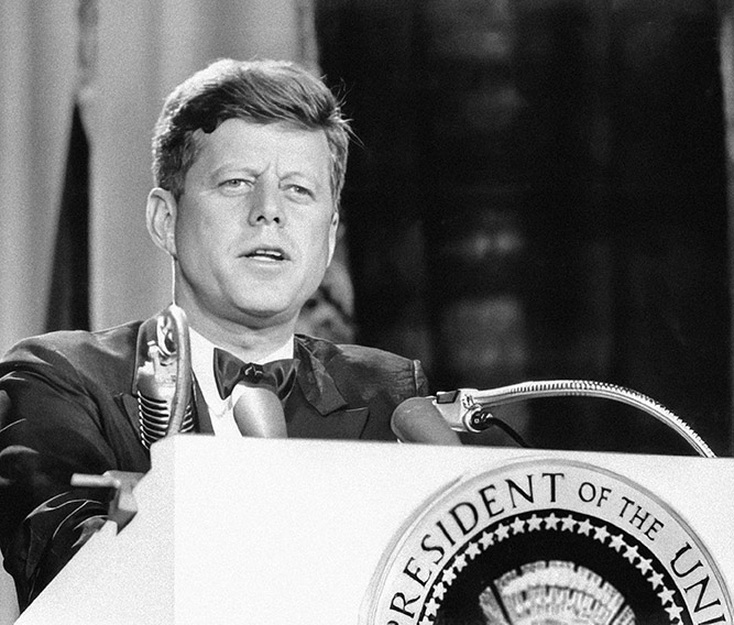 Photo of JFK standing at a podium with a "President of the United States" seal on the front
