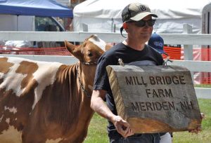 Christian Forman holding a sign "Mill Bridge Farm, Meriden, NH" followed by a cow