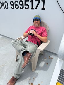 Brian Hoover with binoculars, sitting in a chair on the deck of a boat in the arctic.